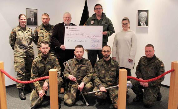 Oberst Lars Persikowski und Oberstleutnant a.D. Martin Heuwinkel präsentieren mit der Küchencrew und weiteren Helfenden des Infostands den symbolischen Spendenscheck. 
